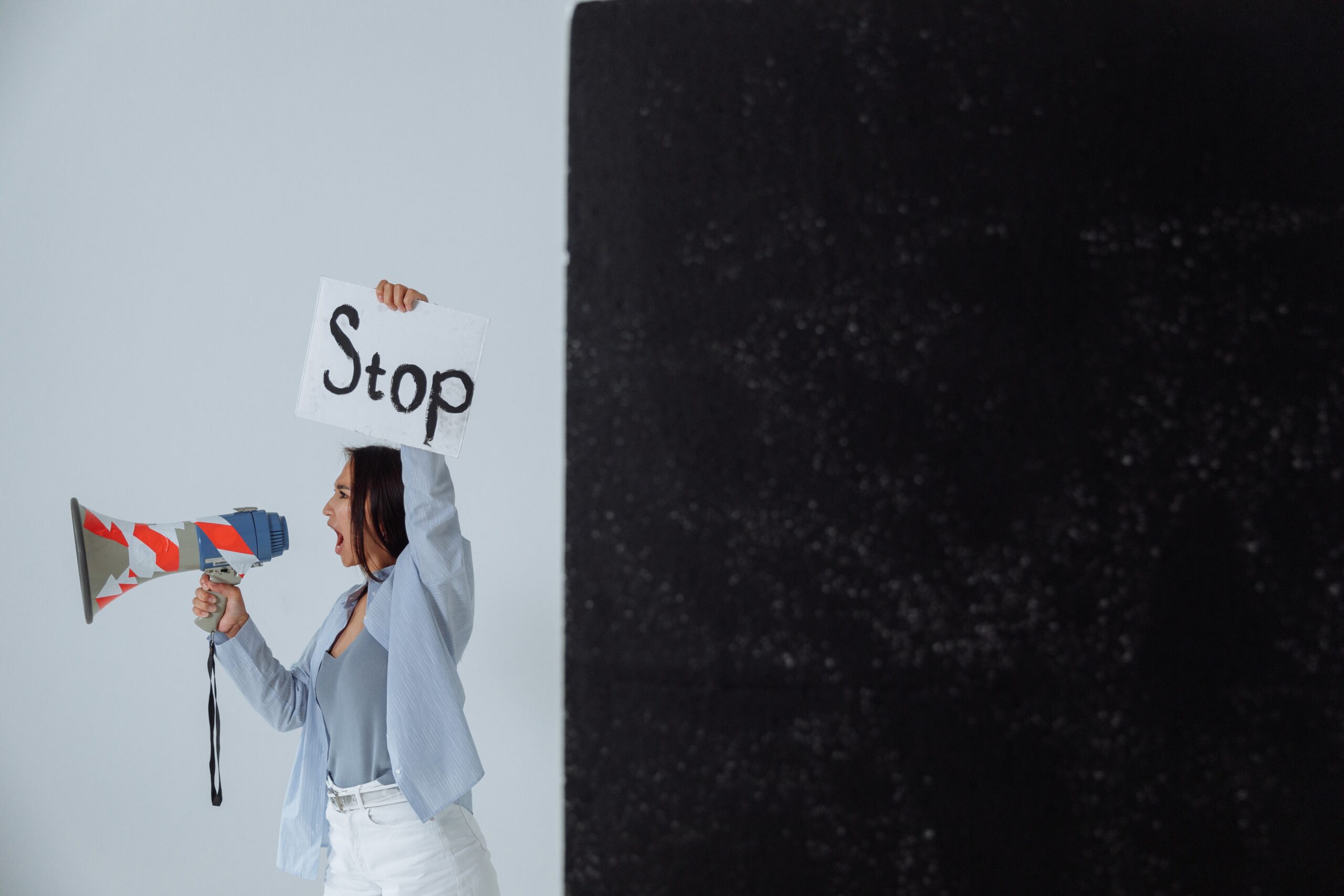 fotografia mostra mulher levantando cartaz com o texto "Stop" com uma das mãos, enquanto utiliza a outra mão para segurar um megafone utilizado para ampliar o alcance de sua voz