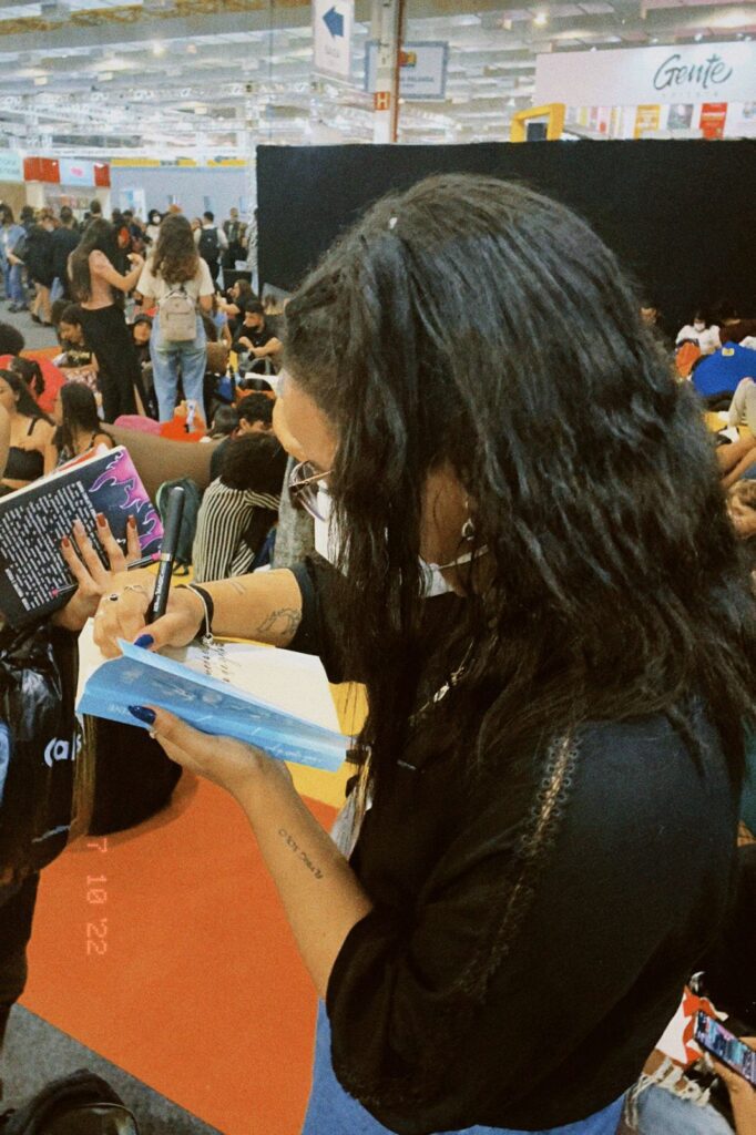 Mulher de cabelos longos e pretos autografa livro. Há várias pessoas ao fundo no que parece ser um evento cultural.