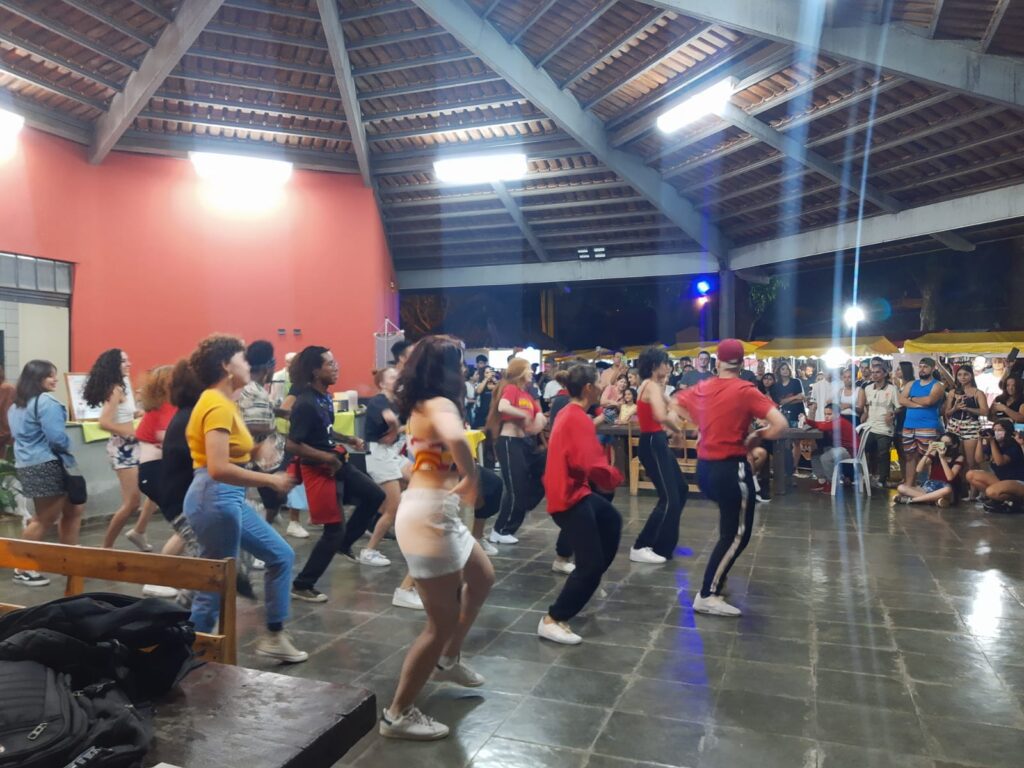 Salão com dançarinas no meio se apresentando e o público no entorno do espaço prestigiando-as.