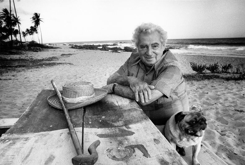 Na foto, o escritor Jorge Amado posa em uma praia com os braços apoiados na mesa, onde está também seu chapéu e sua bengala. No canto da imagem, seu companheiro, um cachorro da raça pug, posa com o escritor