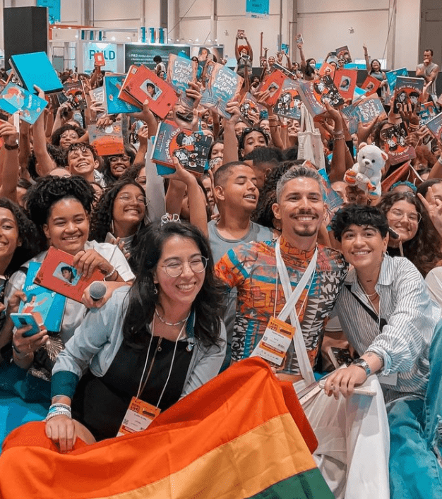 Clara Alves, ao lado de Pedro Rhuas e Elayne Baeta, segurando uma bandeira LGBT, com plateia no fundo