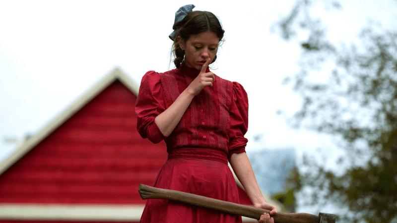 Mia Goth em "Pearl". Mulher branca com o que aparenta ser um celeiro ao fundo, carregando um machado na mão. Seu rosto está sujo de rímel e ela faz um sinal de silêncio.