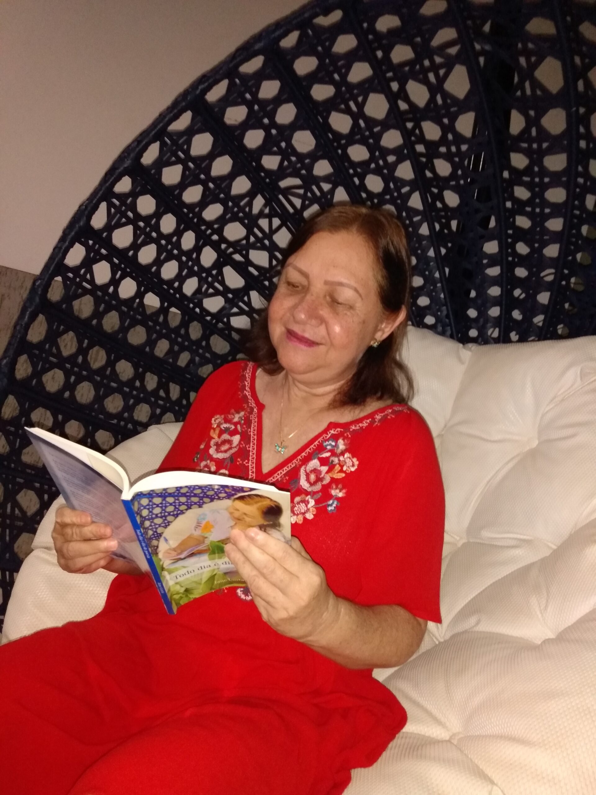 Jane Costa, mulher branca de cabelos curtos e castanhos, usa um vestido vermelho e está sentado em uma poltrona com seu livro "Todo dia é dia de poesia"