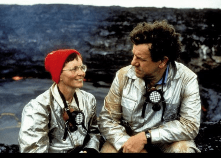 Katia, mulher branca usando óculos e um gorro vermelho, olha sorrindo para Maurice. Os dois usam roupas de proteção e atrás parece ter cinzas vulcanicas.