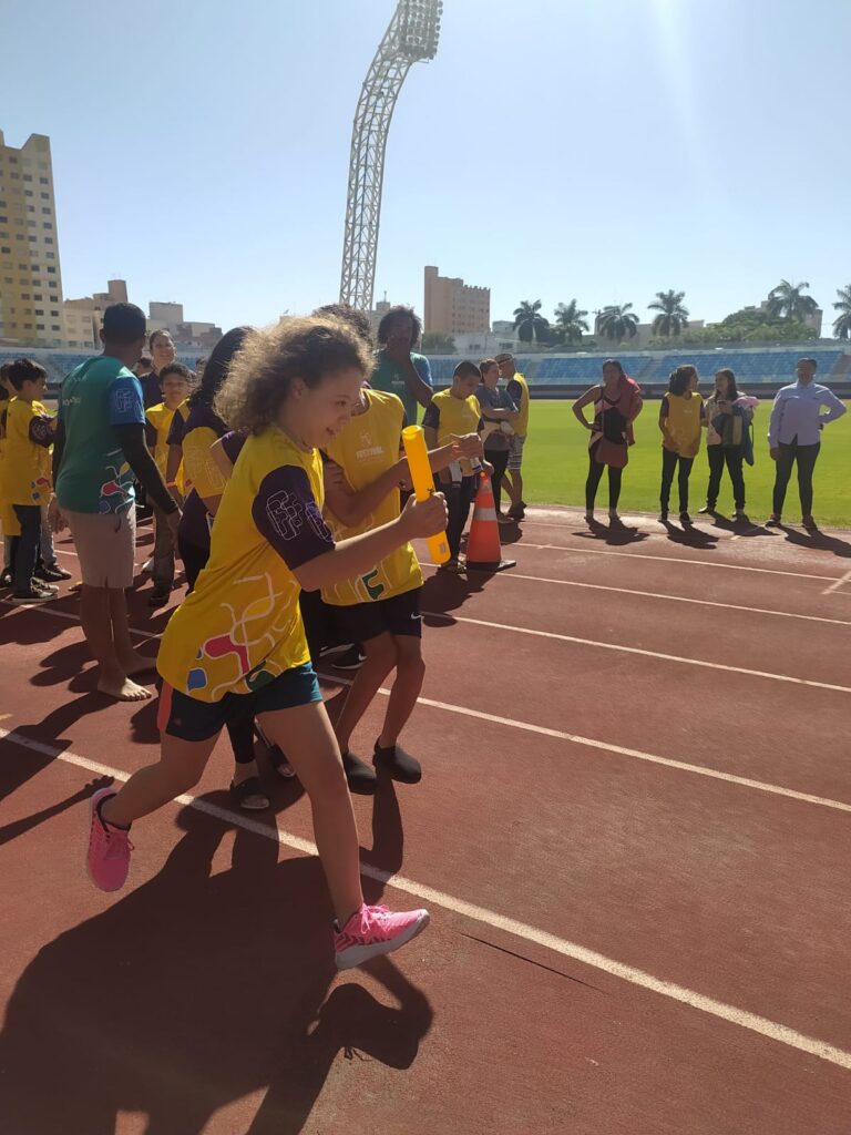 Maria Clara correndo com um bastão na mão em uma pista de corrida. Tem várias pessoas no fundo.
