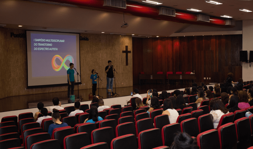 Público e artistas na primeira edição do Simpósio Multidisciplinar do Transtorno do Espectro Autista, em Goiânia. Na foto, costas de pessoas sentadas em cadeiras, de frente para o palco, onde se encontram três pessoas com autismo cantando em apresentação. Ao fundo, a logo e o escrito do 1º Simpósio Multidisciplinar do Transtorno do Espectro Autista. (Foto: Acervo LaTEA/Sandro Henrique Ala)