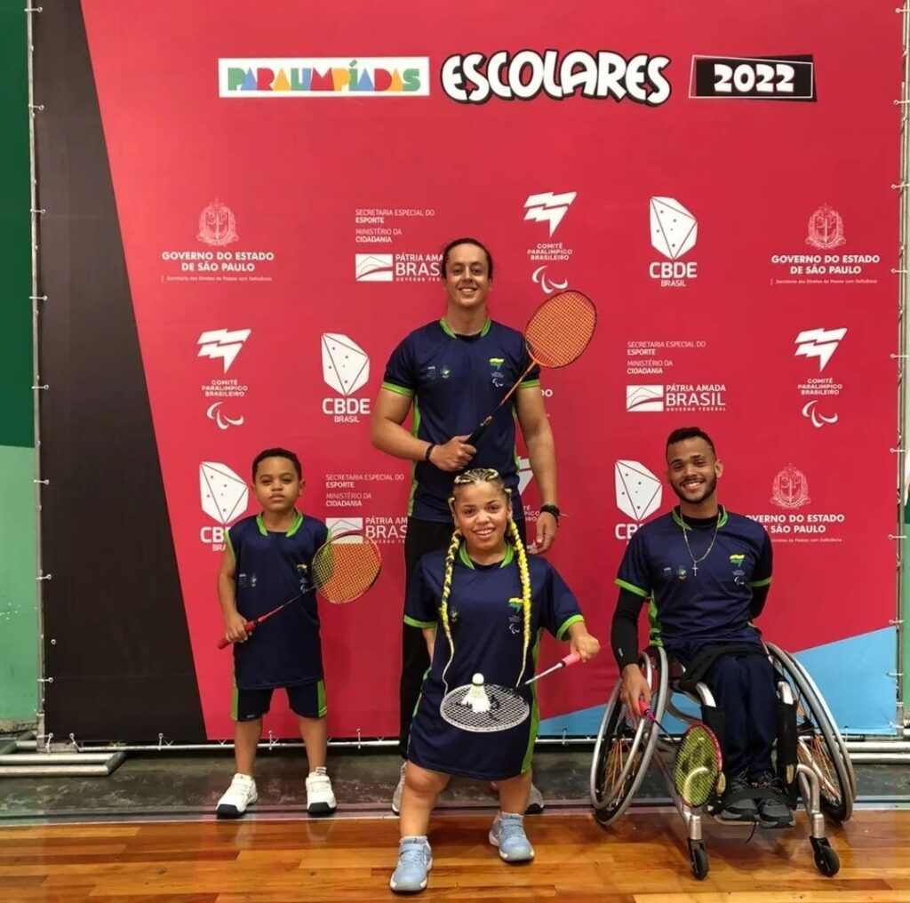 Marcos segurando uma raquete junto a três atletas de parabadminton. No fundo tem um um painel das paralimpíadas escolares 2022.