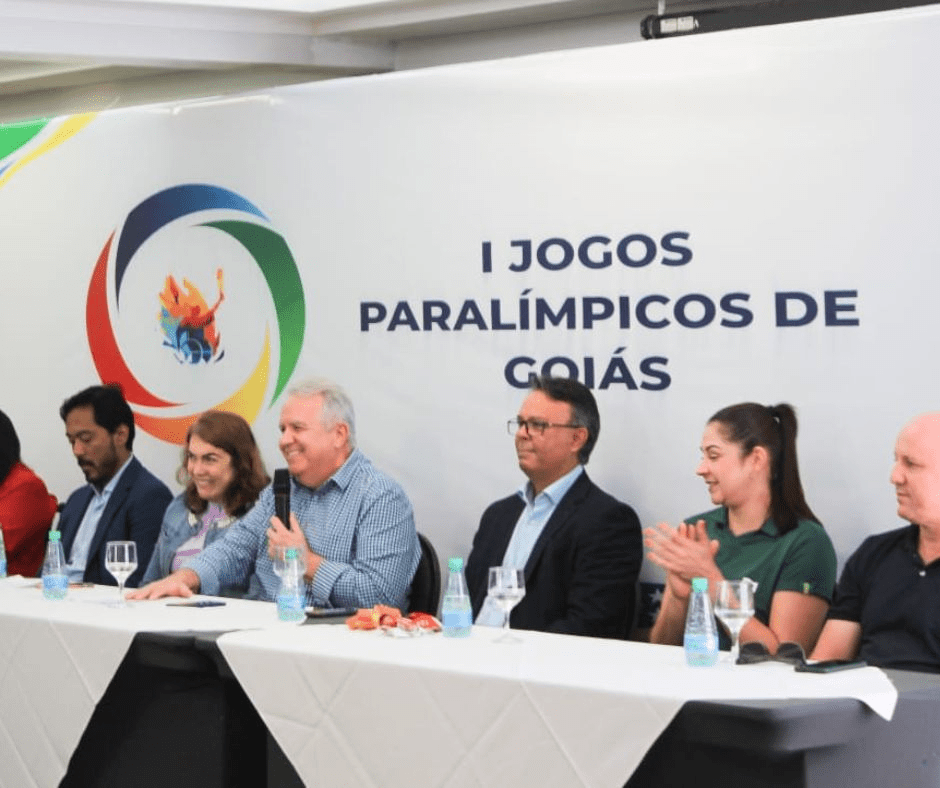 Várias pessoas sentadas na frente do banner dos primeiros jogos paralimpícos de Goiás.