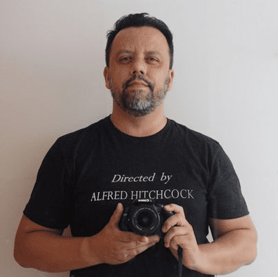 Imagem do Professor Wolney Fernandes. Ele usa uma camiseta escrito "Directed by Alfred Hitchcock" e está segurando uma câmera fotográfica.