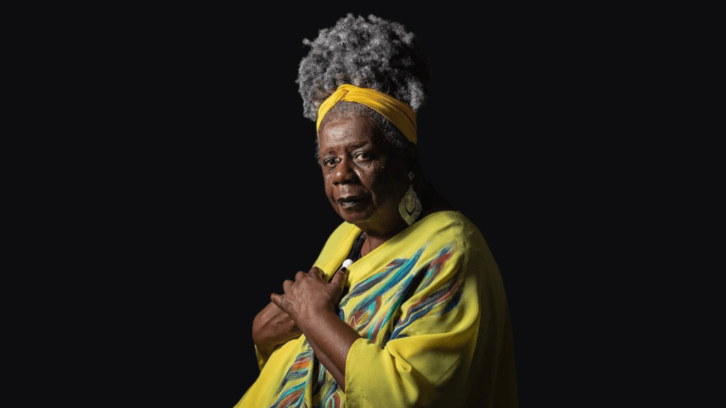Mulher negra e idosa, com vestido amarelo, cabelos amarrados e mãos sobre o peito.