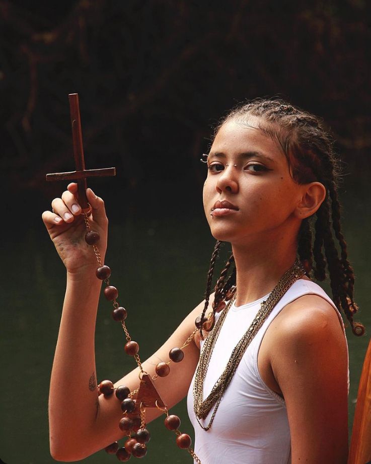 Martinha de Arapo posa para a foto segurando uma cruz de madeira virada de cabeça para baixo.