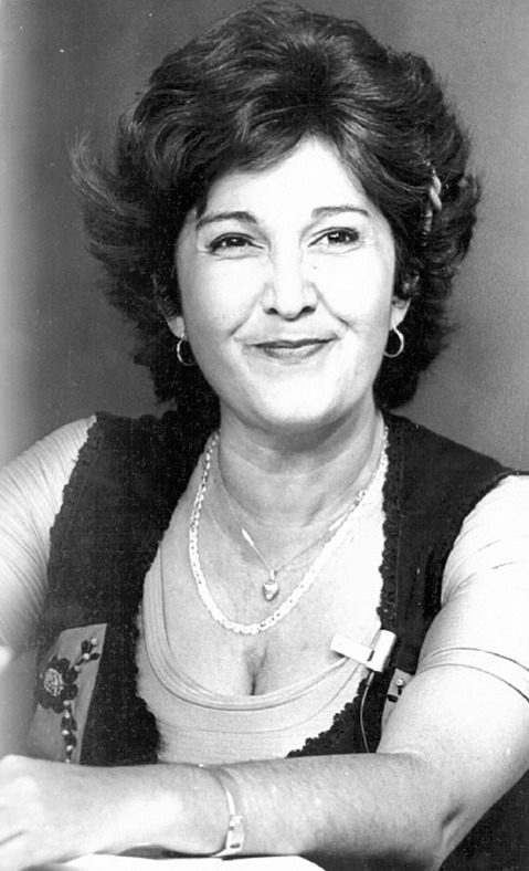 A fotografia em preto e branco mostra uma mulher sorridente em um retrato posado Ela tem cabelos escuros curtos e volumosos penteados para trs e usa brincos de argola pequena e colares finos Sua roupa inclui uma blusa clara de mangas curtas com um colete escuro sobreposto que parece ter detalhes bordadosA mulher tambm usa uma pulseira no pulso esquerdo e tem preso roupa um pequeno microfone de lapela sugerindo que poderia estar participando de uma gravao ou entrevista Seu sorriso discreto e acolhedor e o fundo da imagem liso e neutro destacando seu rosto e expresso serena