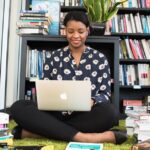 A woman sitting on a carpet in a library working on her laptop, surrounded by books.