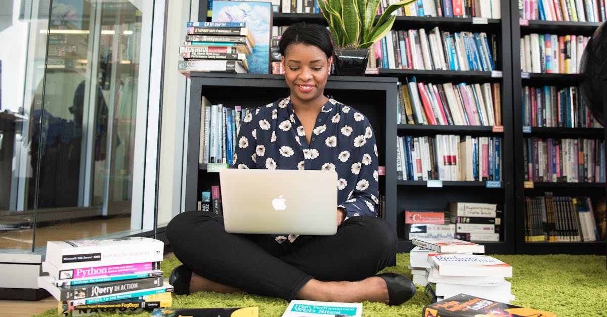 A woman sitting on a carpet in a library working on her laptop, surrounded by books.